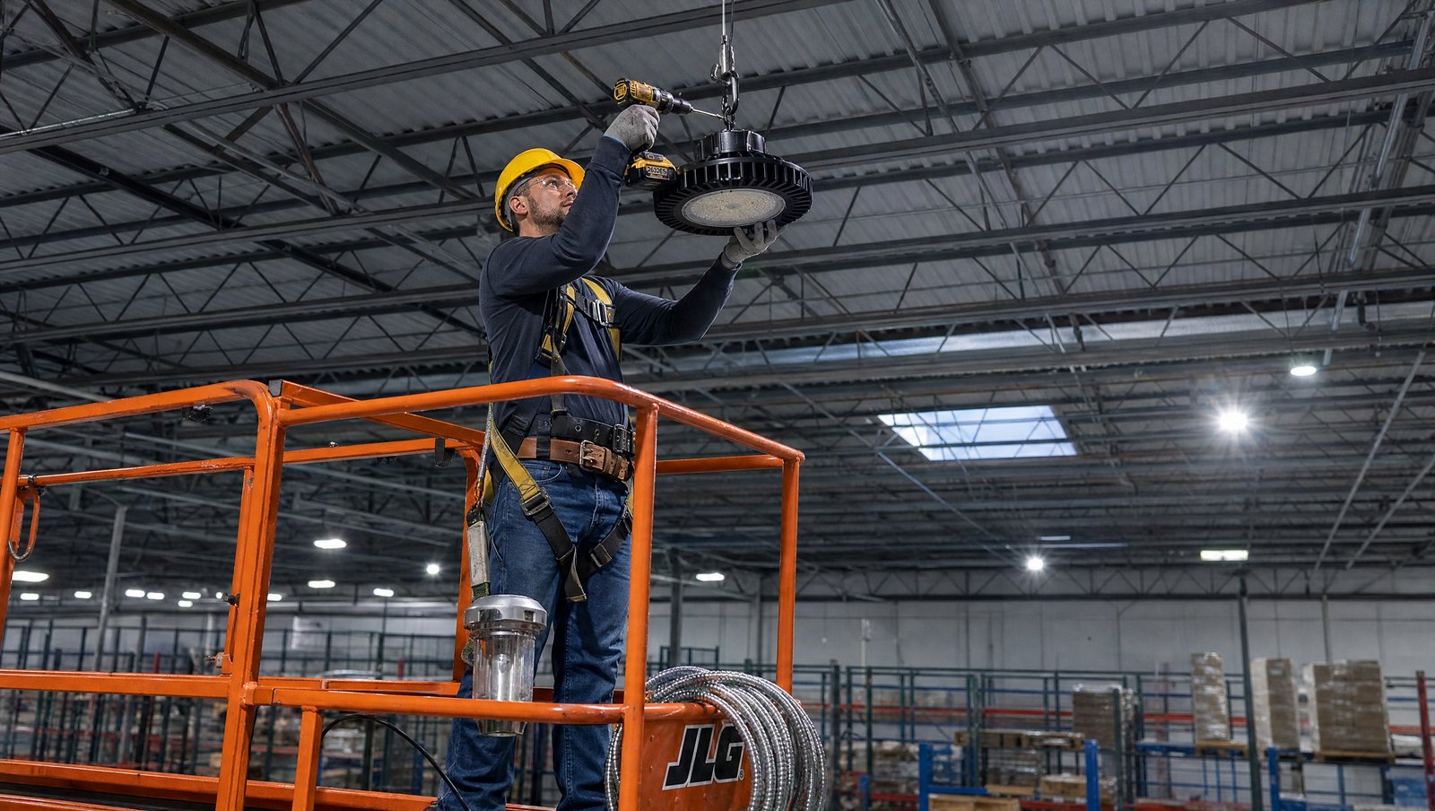Electrical contractor in a yellow hard hat and full body safety harness standing on an orange scissor lift platform installing a round 200W LED UFO high bay fixture to a J-hook on a steel ceiling joist of a warehouse with cordless drill in hand