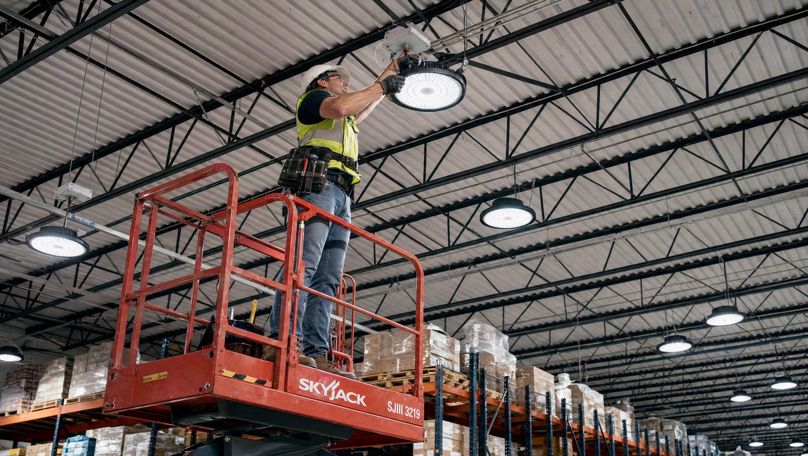 Professional electrician on a scissor lift installing an LED high bay fixture in an active warehouse