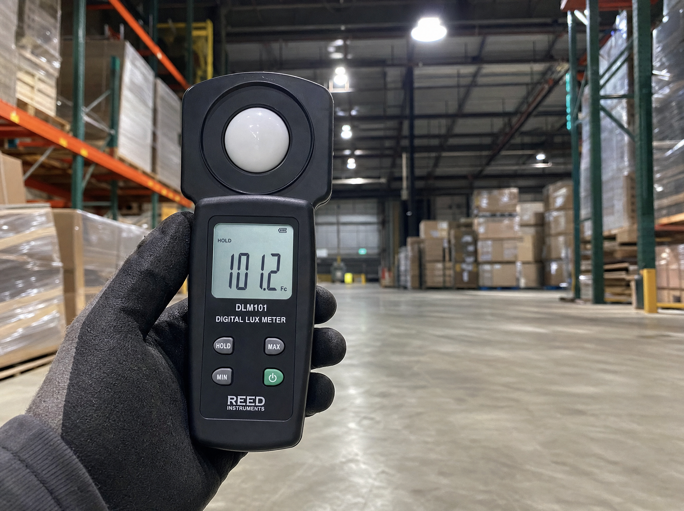 Close-up of a digital lux meter held by a gloved hand at floor level inside a working warehouse with a 200W LED high bay fixture illuminated overhead, the meter screen reading a foot-candle value